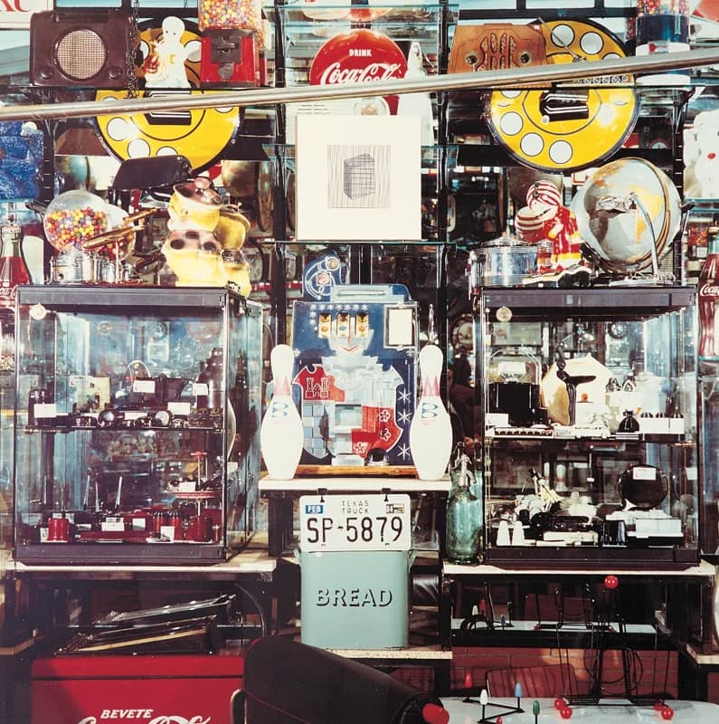 Vetrina stipata di oggetti vintage e giocattoli, con due grandi dischi gialli, una macchina da gioco, teche di vetro, insegne Coca-Cola e un mappamondo, nei toni del rosso, giallo, nero e argento.