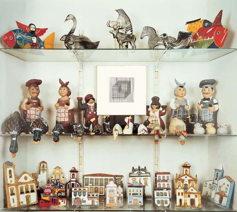 Tre ripiani di vetro espongono figurine e sagome dipinte: pesci, uccelli e zebre in alto, personaggi e animali al centro, piccole case e chiese in basso. Al centro del ripiano mediano è appesa una cornice bianca con un disegno geometrico grigio e nero.