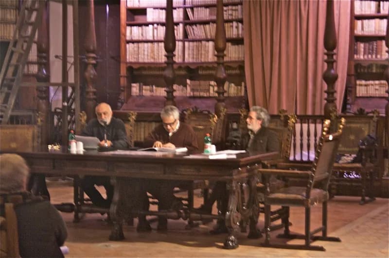 Tre uomini siedono a un grande tavolo di legno scuro, con carte e bottiglie d’acqua davanti, in una biblioteca dalle alte scaffalature colme di libri, tende rosa e arredi intagliati.