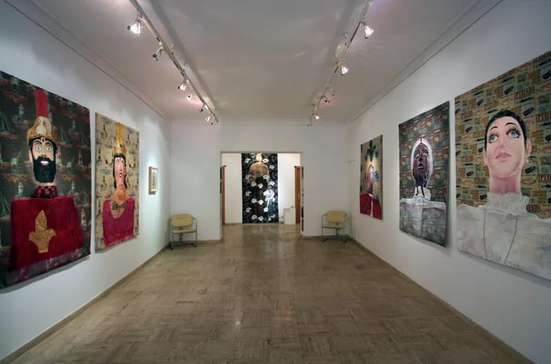 Veduta di una galleria dalle pareti bianche, con grandi tele raffiguranti volti e figure stilizzate appese su entrambi i lati; in fondo, attraverso una porta, si vede un’opera scura con motivi floreali bianchi.