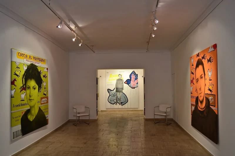 Veduta di una galleria dalle pareti bianche, con due grandi ritratti su fondi giallo e arancione alle pareti laterali e, in fondo, un’opera con figure umane e animali stilizzate; due sedie metalliche fiancheggiano il passaggio.